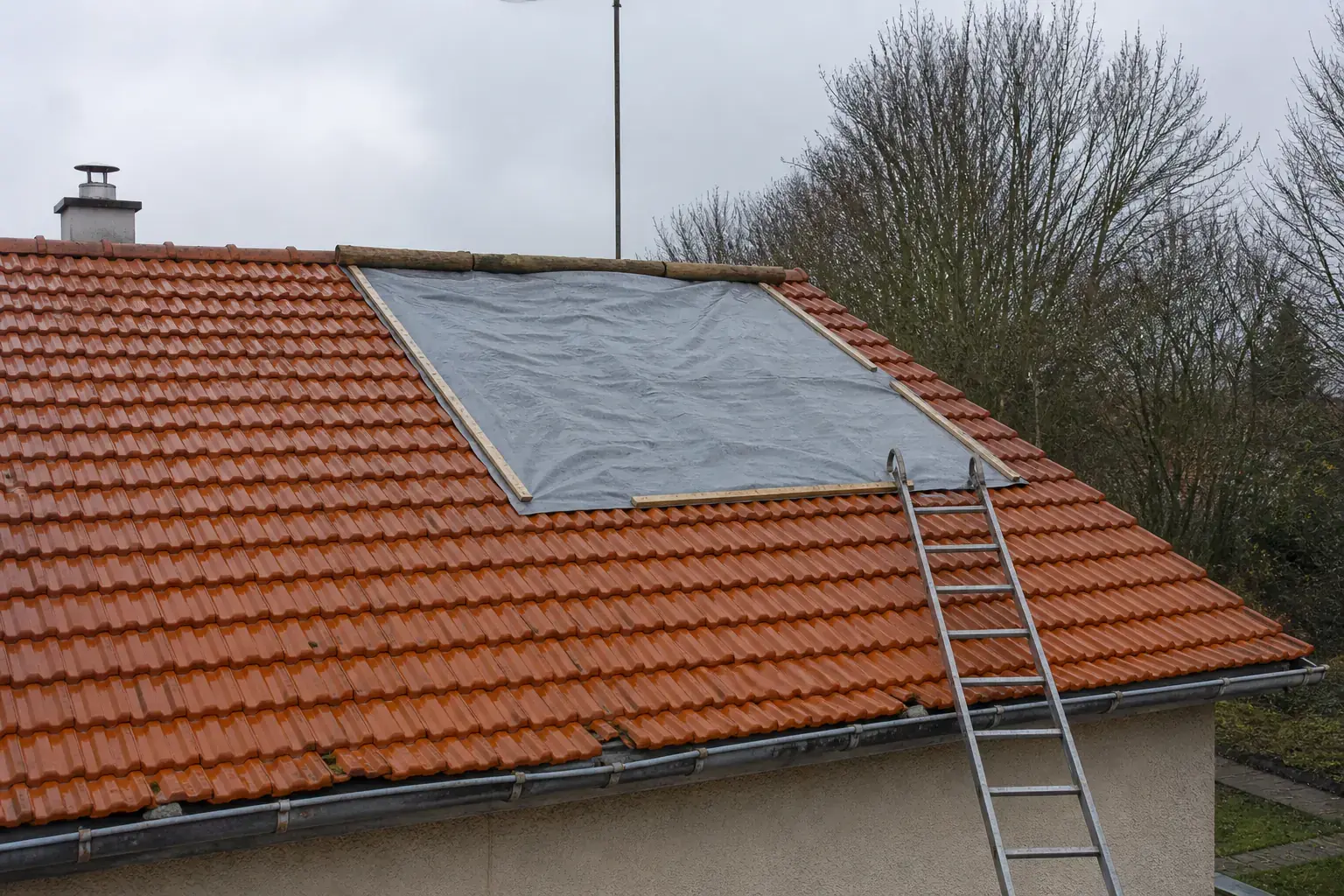 Toit de tuiles mécaniques mouillé après une tempête : une bâche grise tenue par des lattes de bois couvre une partie du versant, une échelle est appuyée contre le toit