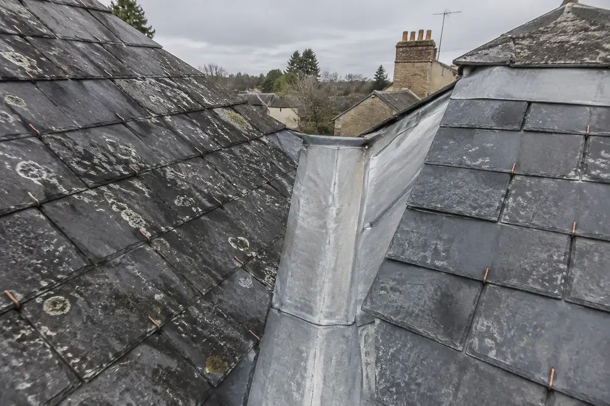 Noue en zinc entre deux pans de toiture en ardoise naturelle, crochets de cuivre visibles, par ciel couvert