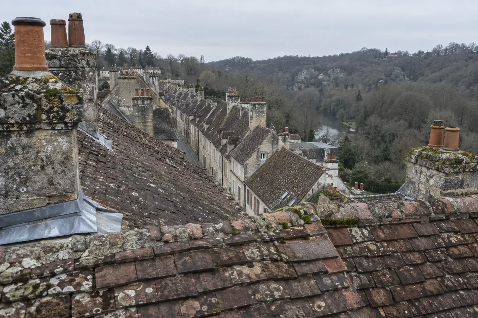 Rangée de toits de tuiles alignés le long d’une rue droite, souches de cheminée en pierre, vallée boisée en contrebas, par ciel couvert