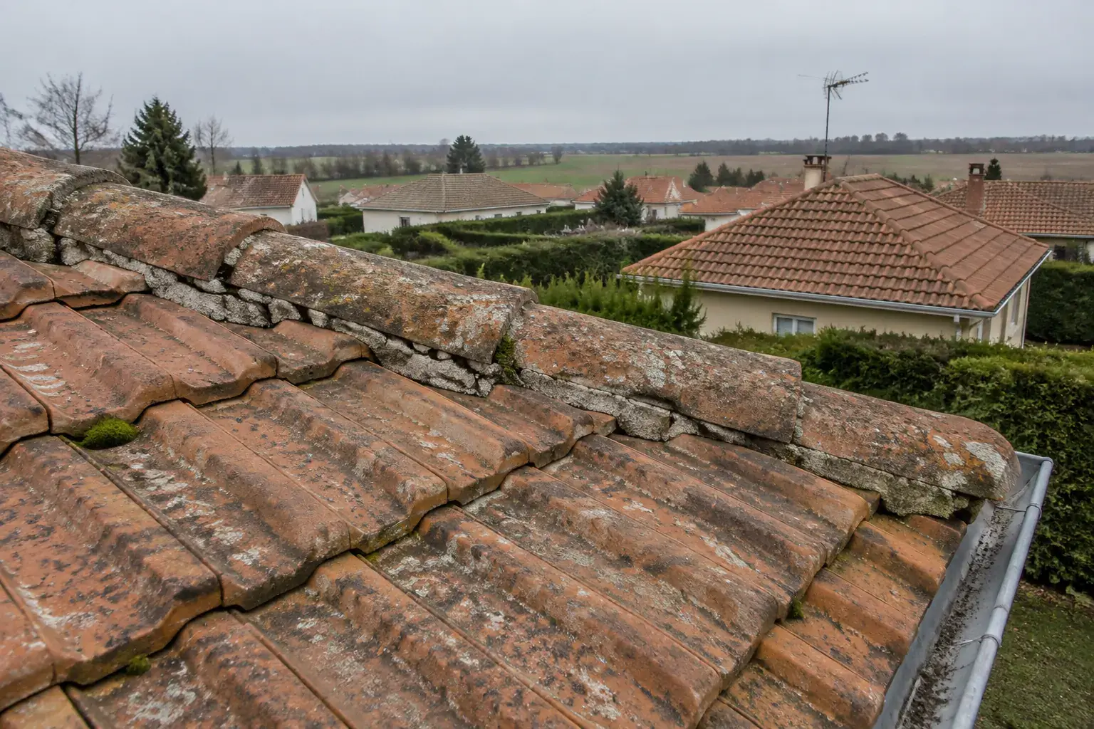 Faîtage scellé au mortier sur un toit de tuiles mécaniques de pavillon, autres pavillons et champs à l’horizon, par ciel couvert
