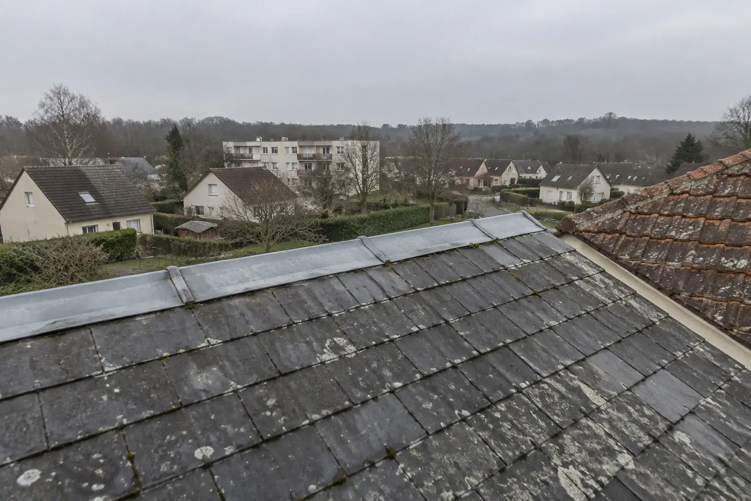 Toit d’ardoise artificielle avec faîtage en zinc sur un pavillon, quartier résidentiel et vallée boisée au fond, par ciel couvert